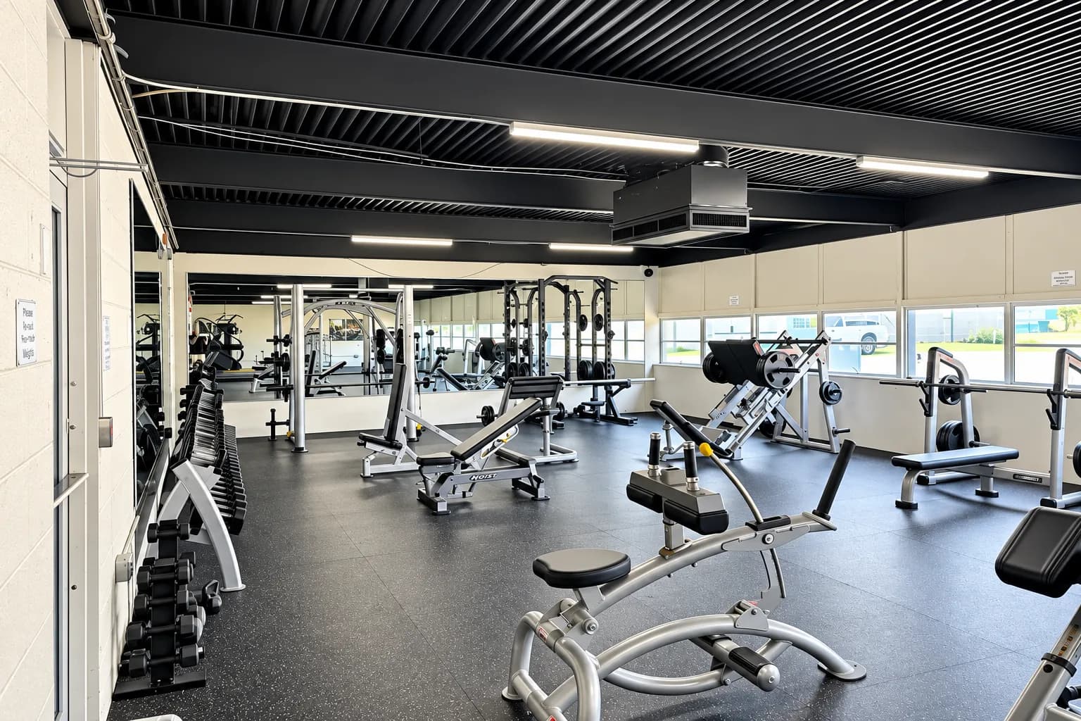 The Fitness Zone: weight benches, racks, and machines under a black ceiling with windows along one wall